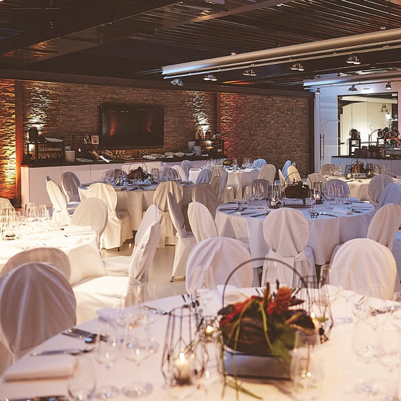 Ein Bankettsaal mit runden Tischen mit weißen Tischdecken und Stuhlhussen, geschmückt mit einfachen Blumengestecken, umgeben von hübsch angeordneten Weingläsern und Besteck, schafft einen eleganten Ort für jede Veranstaltung.