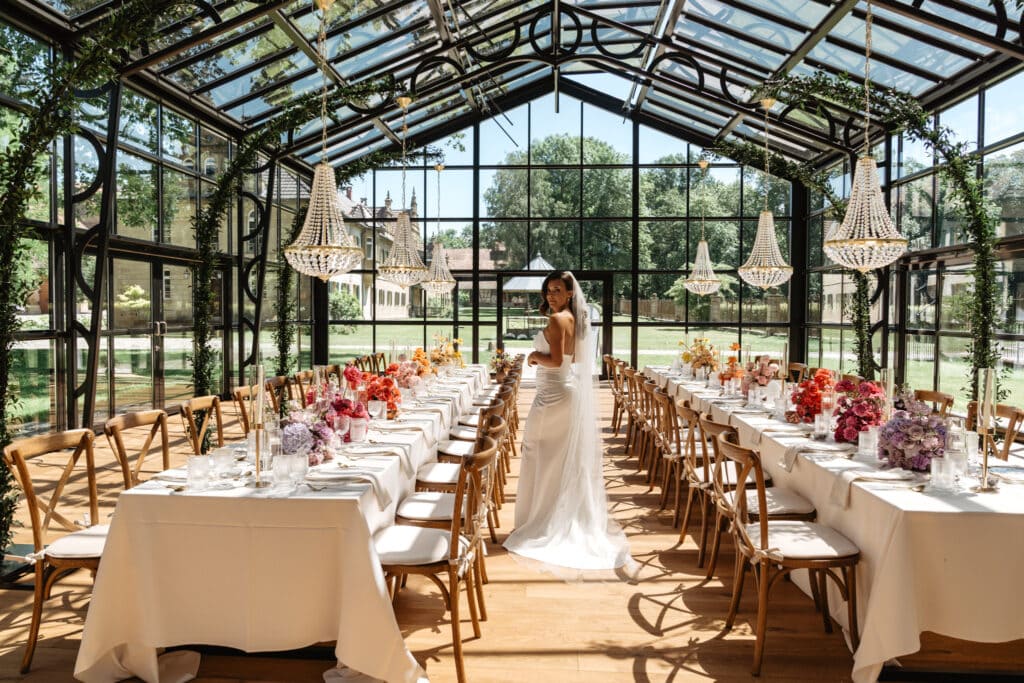 Eine Person im Brautkleid steht in der Mitte einer wunderschön dekorierten, verglasten Halle mit Kronleuchtern, langen Esstischen und floralen Tischdekorationen.