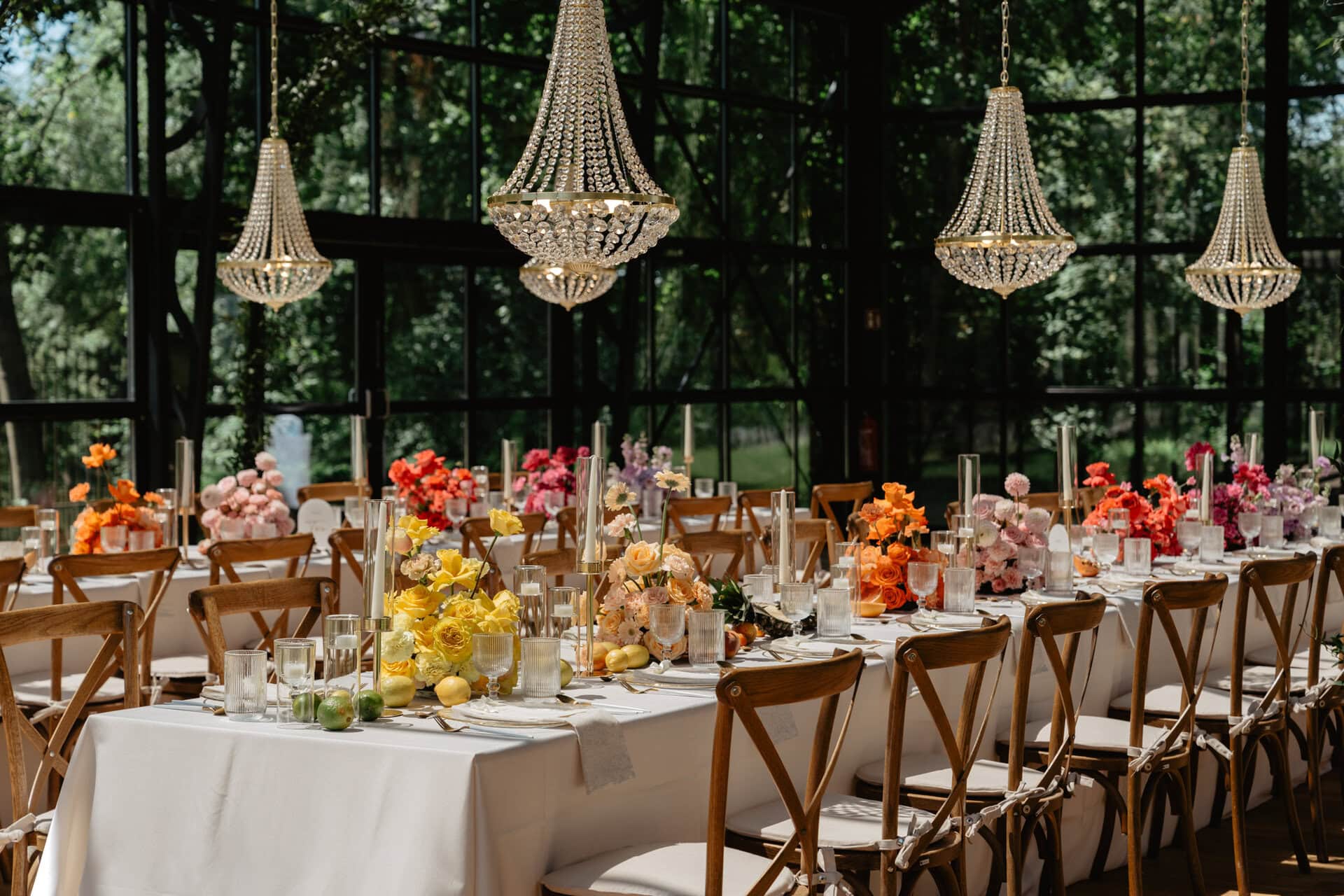 Ein langer Banketttisch mit weißer Tischdecke, geschmückt mit farbenfrohen Blumenarrangements und darüber liegenden Kristallleuchtern, in einem Raum mit großen Fenstern und Blick auf die Bäume draußen.