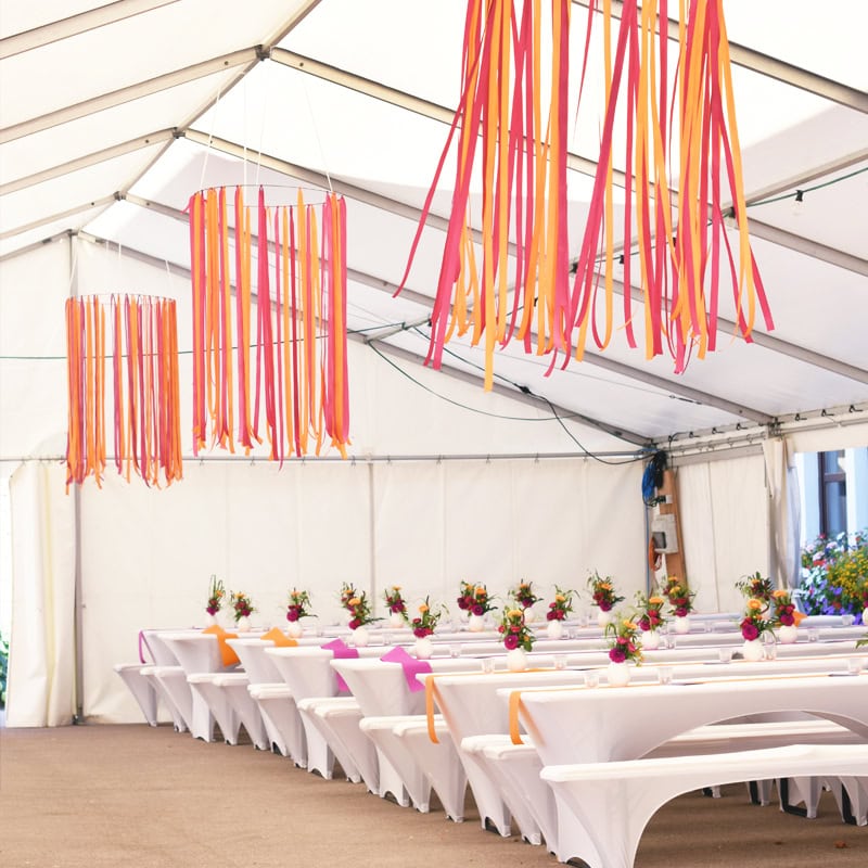 Es ist ein großes weißes Zelt mit Reihen weißer Tische und Bänke aufgebaut, das mit bunten Blumen und hängenden orangefarbenen und rosa Luftschlangen dekoriert ist.