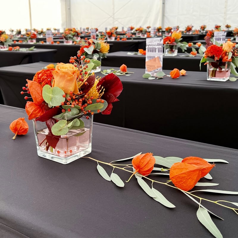 Ein großer Raum mit langen Tischen, die mit schwarzen Tischdecken bedeckt sind. Jeder Tisch hat Glasaufsätze mit orangefarbenen, roten und gelben Blumen und verstreuten Blättern als Dekoration.